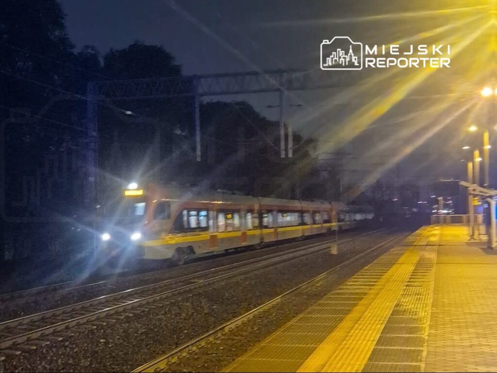 Pociąg z oświetleniem nocnym zbliża się do stacji, gdzie widoczna jest platforma z oznaczeniami.