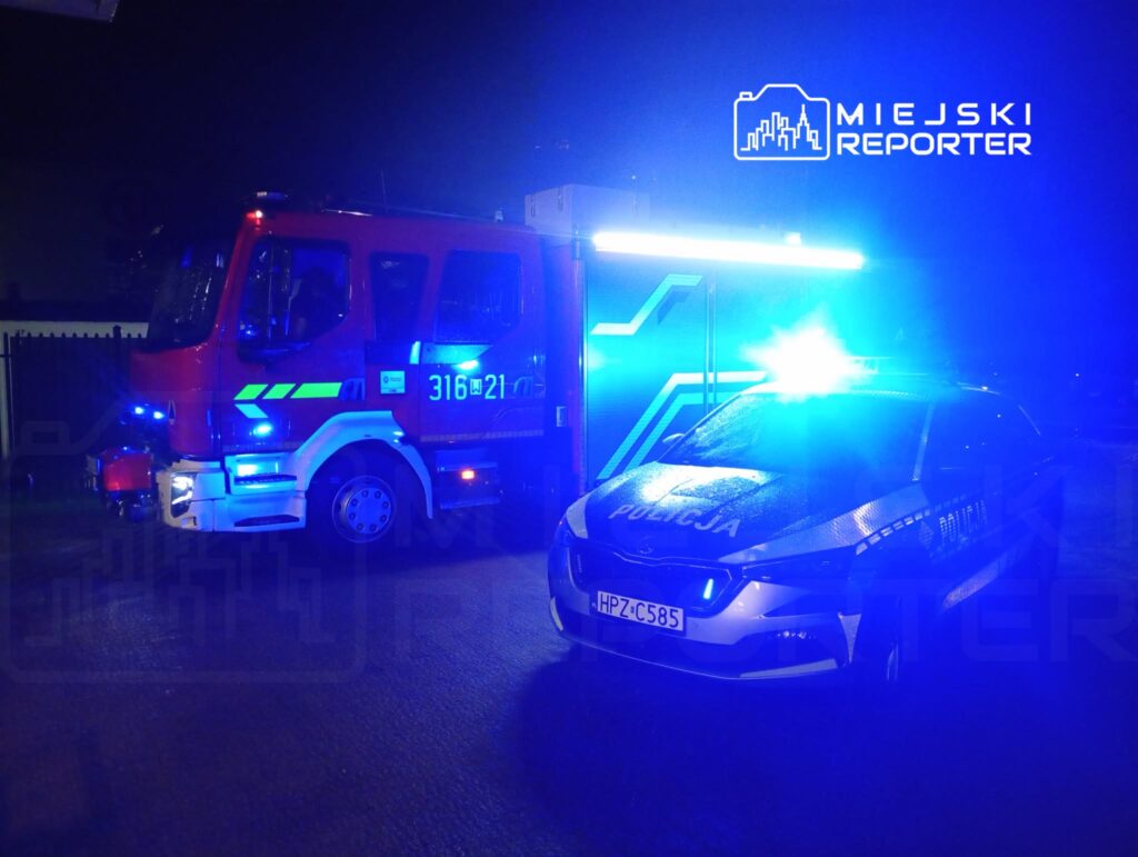 W nocy pojazdy ratunkowe, w tym wóz strażacki i radiowóz policyjny, zaparkowane na oświetlonej ulicy, emitują niebieskie światła.