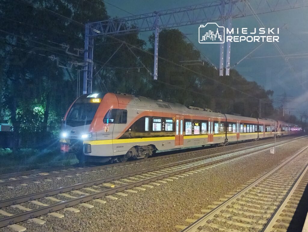 Pociąg osobowy zatrzymany na stacji w nocy, oświetlony reflektorami, w otoczeniu drzew i torów kolejowych.