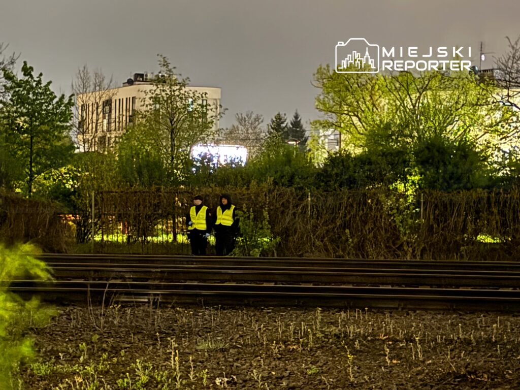 Dwaj mężczyźni w odblaskowych kamizelkach stoją obok torów kolejowych w oświetlonym terenie zielonym.