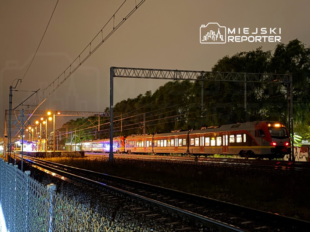 Pociąg pasażerski zatrzymany na stacji w oświetlonym nocą otoczeniu z widocznymi torami i latarniami.