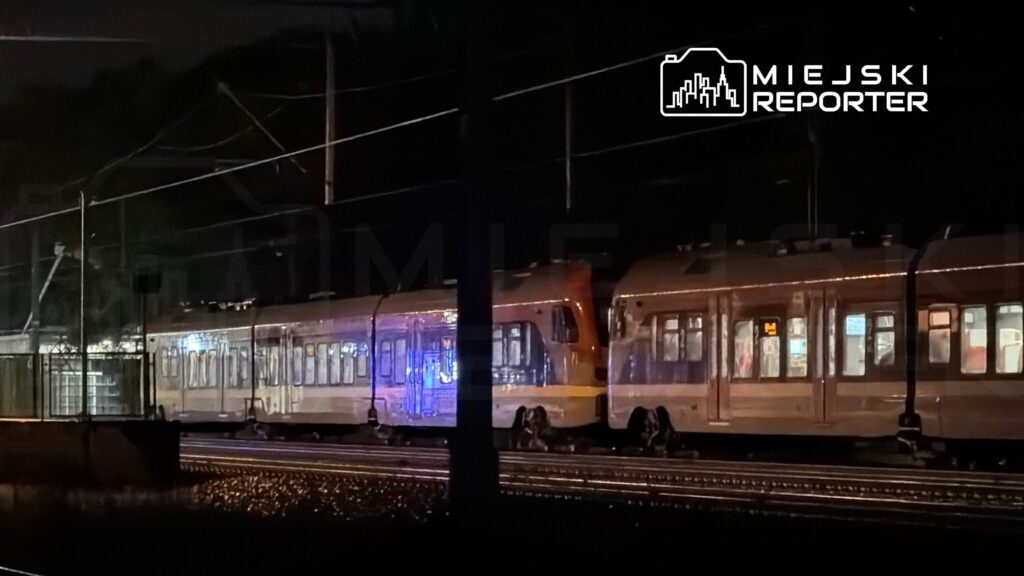 Pociąg tramwajowy zatrzymany na stacji w nocnym otoczeniu, oświetlony niebieskimi światłami.