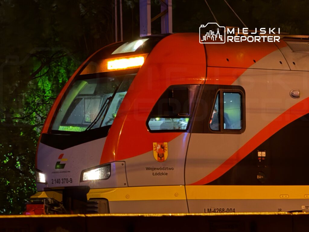 Pociąg elektryczny z logo województwa łódzkiego zatrzymany na stacji w nocy, oświetlony zielonymi światłami.