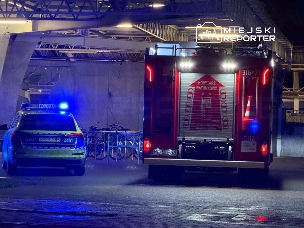 Policyjny radiowóz z włączonymi sygnałami świetlnymi oraz wóz strażacki z widocznym znakiem "KORYTARZ RATUNKOWY" na nocnym tle.
