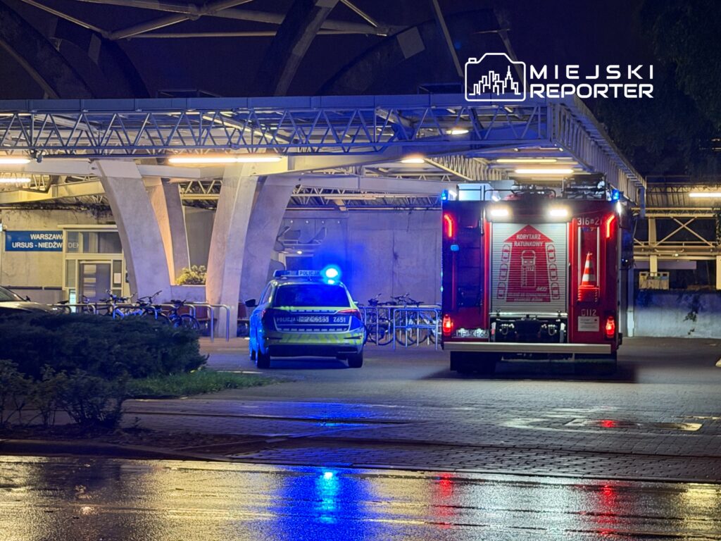 Policyjny radiowóz i wóz strażacki zaparkowane w oświetlonym miejscu przed budynkiem, w tle rowery.