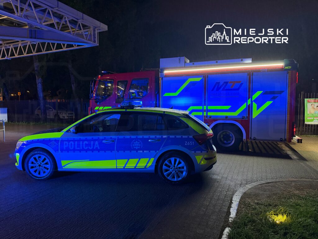 Policyjny radiowóz z niebieskimi i żółtymi oznaczeniami stoi obok wozu strażackiego na oświetlonym terenie nocą.