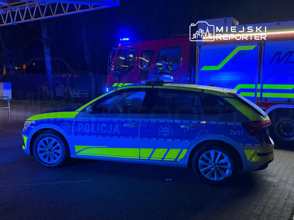 Policyjny radiowóz z oznaczeniem "POLICJA" zaparkowany w pobliżu wozu strażackiego, otoczenie oświetlone niebieskimi światłami.