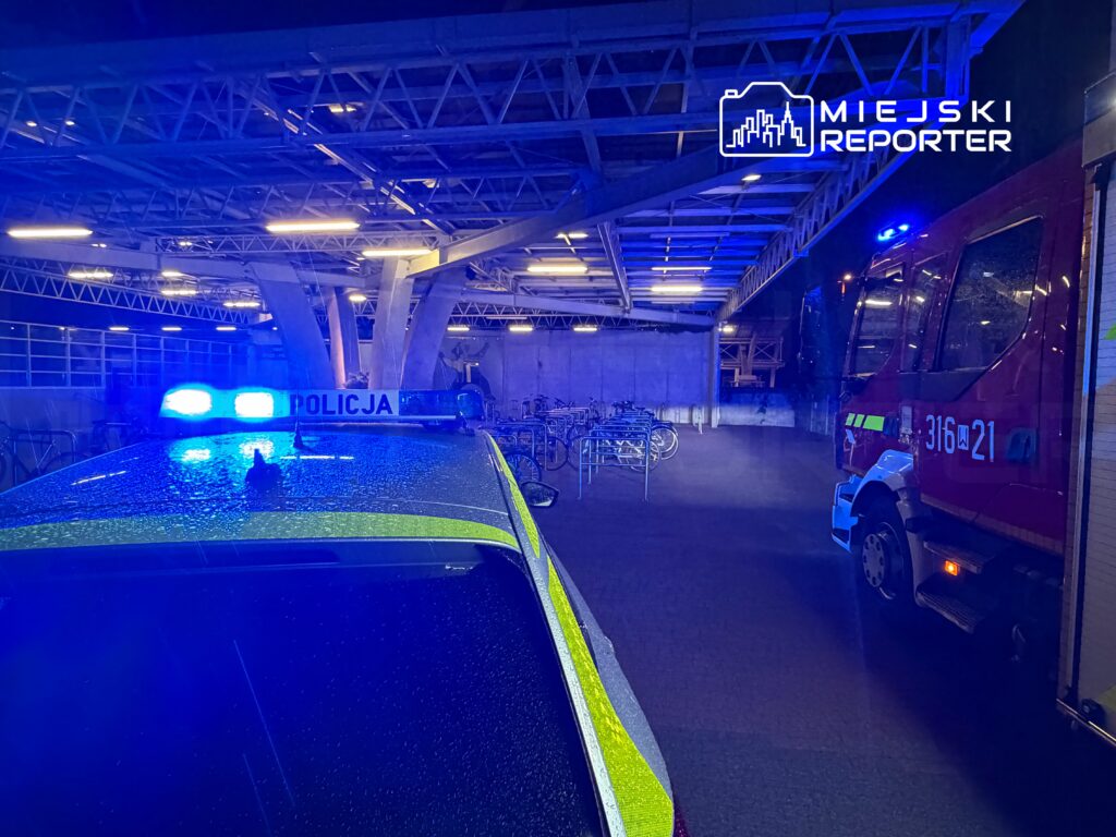 Radiowóz policyjny z włączonymi sygnałami świetlnymi stoi obok wozu strażackiego w oświetlonym garażu.