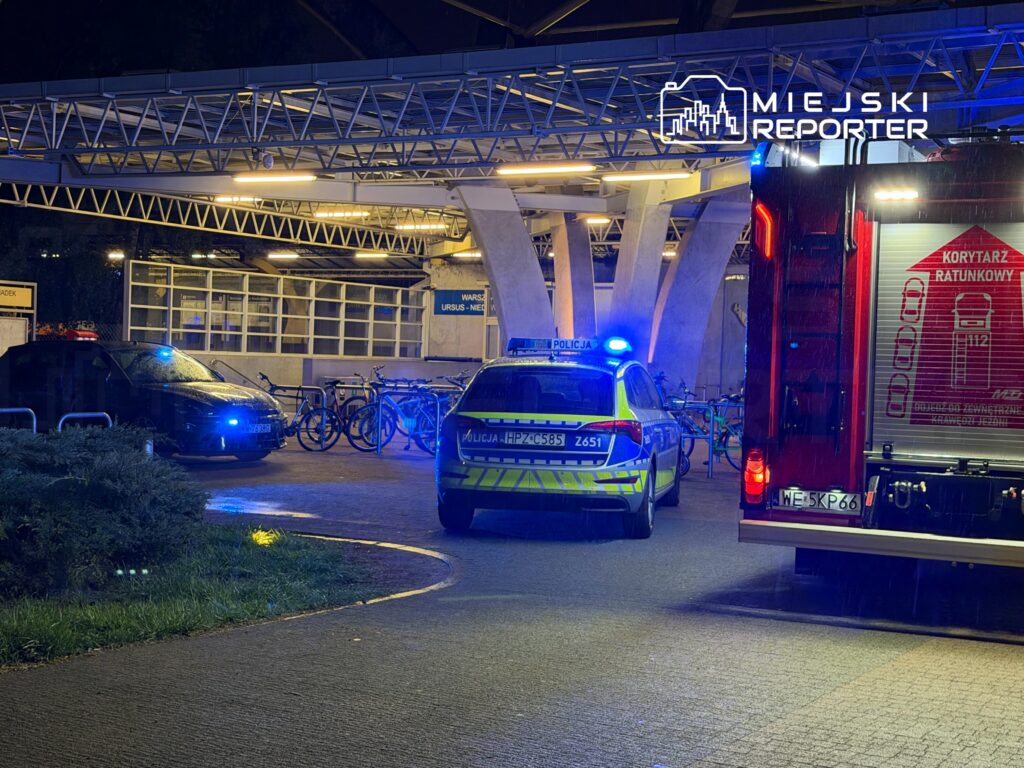 Policyjny radiowóz oraz wóz strażacki z włączonymi sygnałami świetlnymi w pobliżu stacji rowerów miejskich.