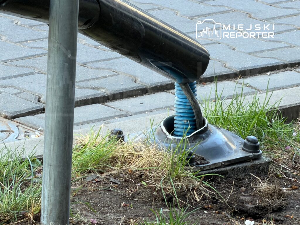 Czarna rura z niebieską sprężyną podłączona do metalowego słupa, osadzona na trawniku obok kostki brukowej.