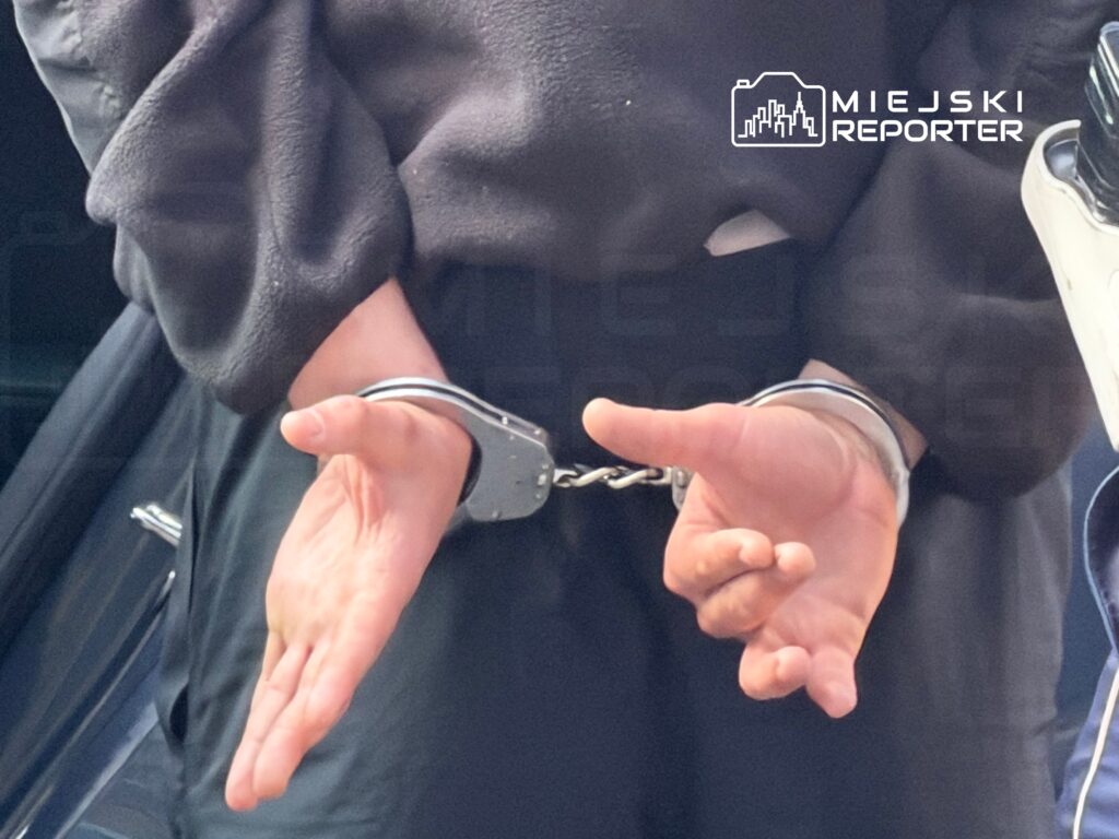 Mężczyzna z rękami skuty w kajdankach stoi obok pojazdu policyjnego.