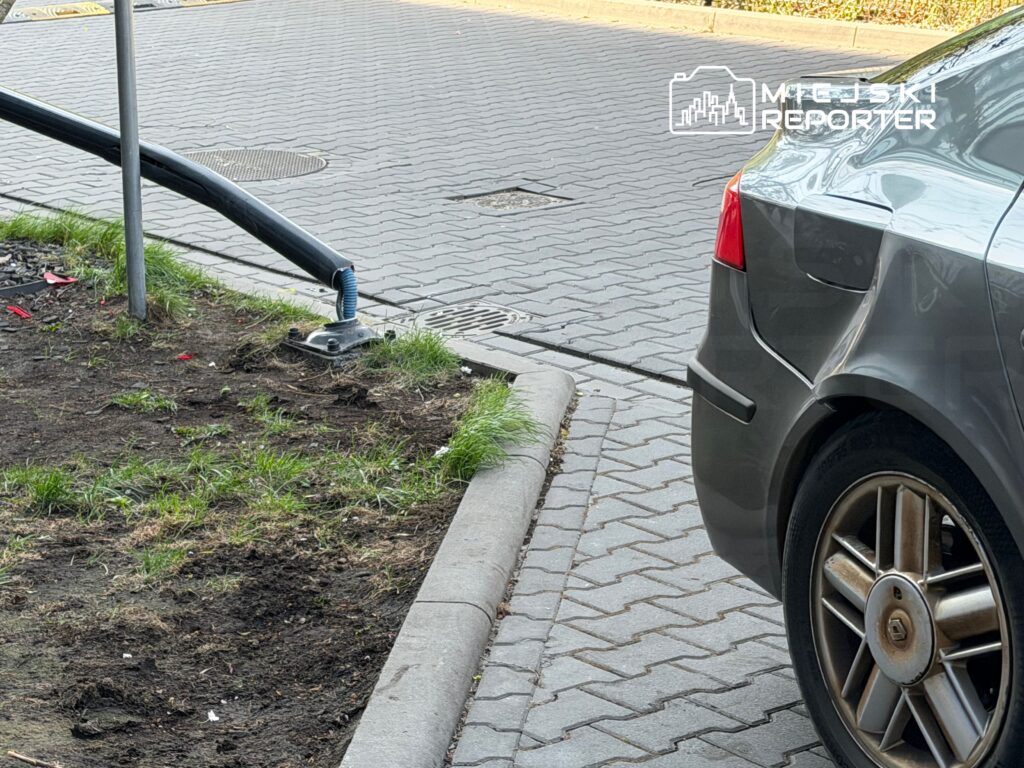 Samochód zaparkowany na chodniku obok metalowego słupa z elastycznym przewodem, w otoczeniu trawnika i kostki brukowej.