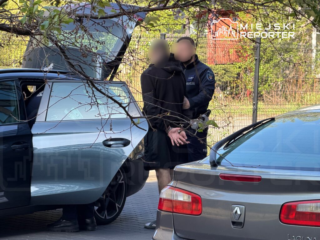 Mężczyzna w rękach policji stoi obok otwartego samochodu, podczas gdy drugi funkcjonariusz obserwuje sytuację.