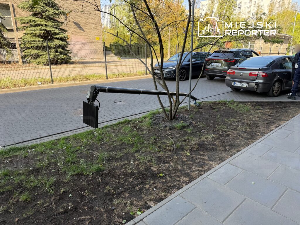 Przydrożny słup z zamontowanym panelem słonecznym, obok zaparkowane samochody na utwardzonej nawierzchni.