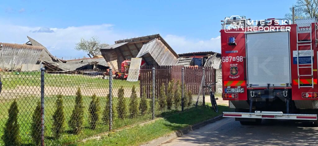 Zniszczony budynek gospodarczy z usuniętym dachem na tle zielonej trawy, obok stoi wóz strażacki.