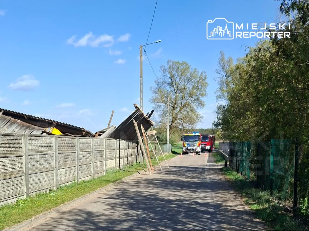 Zniszczony dach drewnianego budynku oparty na ogrodzeniu, obok widoczny wóz strażacki na drodze.