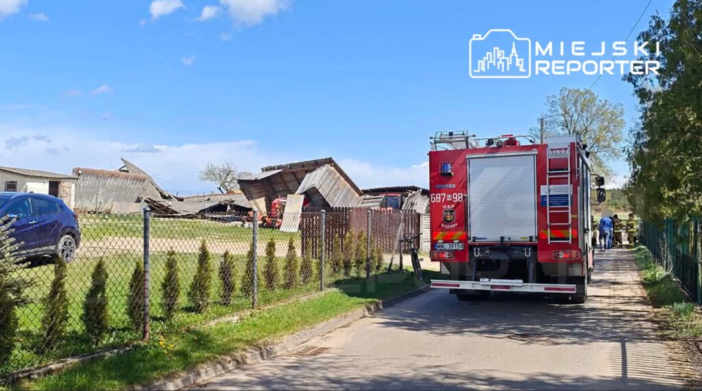 W tle zniszczony budynek gospodarczy, a z przodu woz strażacki zaparkowany przy ogrodzeniu, obok strażacy rozmawiają.