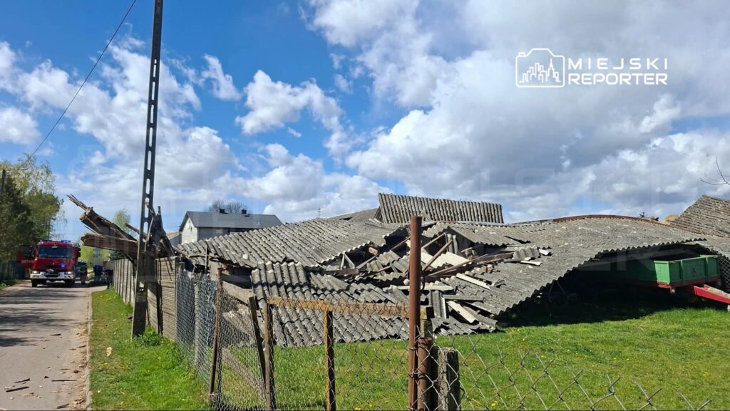 Zniszczony dach budynku gospodarczego leży na ziemi, obok stoi wóz strażacki na tle niebieskiego nieba z chmurami.