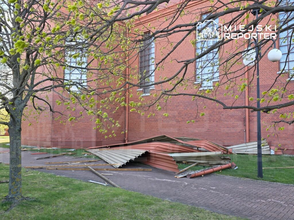 Zniszczona konstrukcja dachowa leży na trawniku obok czerwonej ceglanej ściany budynku, otoczona gałęziami drzewa.