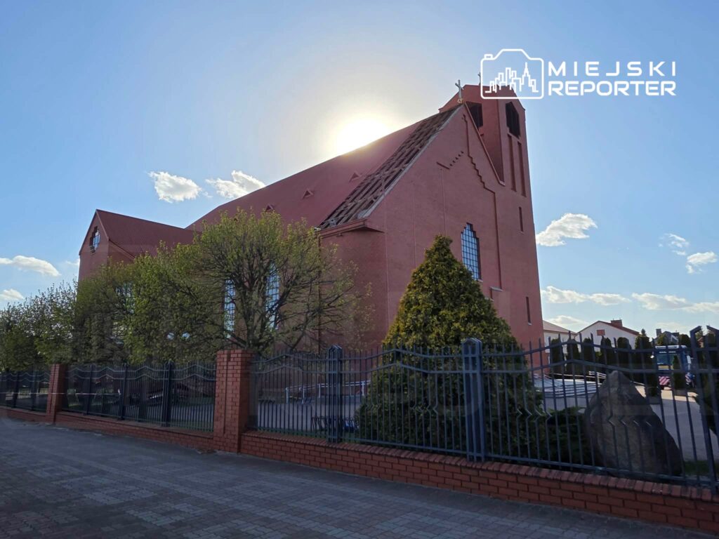 Czerwony kościół z dużymi oknami, otoczony zielonymi drzewami i ogrodzeniem, na tle niebieskiego nieba.