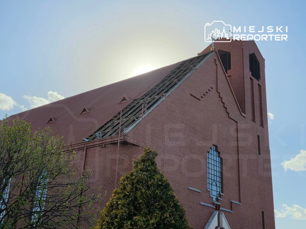 Czerwony kościół z widocznym uszkodzonym dachem, w tle słońce za chmurami, otoczony zielenią drzew.
