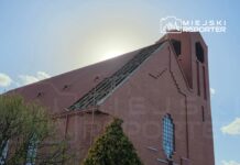 Czerwony kościół z widocznym uszkodzonym dachem, w tle słońce za chmurami, otoczony zielenią drzew.