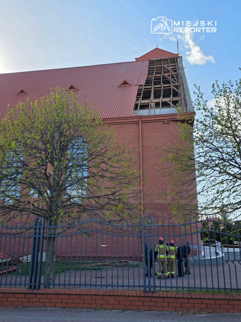 Grupa strażaków w mundurach omawia sytuację przy budynku z czerwoną cegłą i uszkodzoną dachówką.