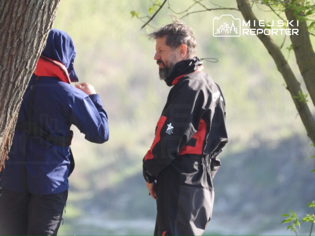 Dwaj mężczyźni w odzieży roboczej rozmawiają na brzegu rzeki, w tle widoczne drzewa i zieleń.