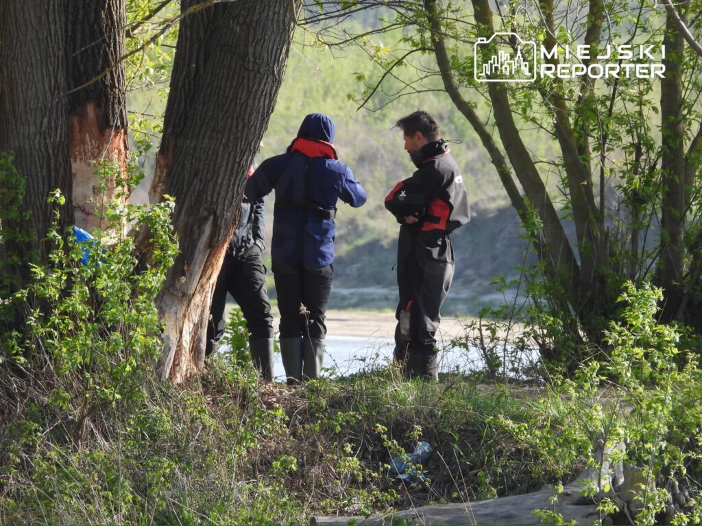 Grupa trzech osób w odzieży roboczej dyskutuje w pobliżu rzeki, otoczona zielonymi drzewami i krzewami.