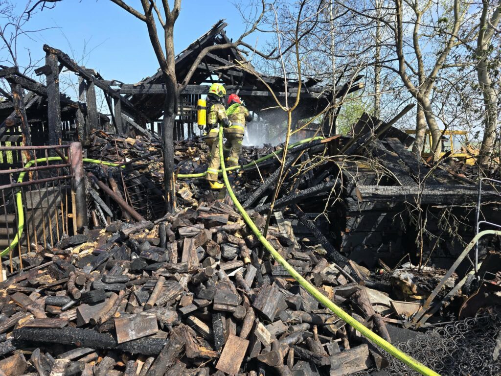 Strażacy gaszą pożar w zniszczonym budynku, otoczonym stosami spalonego drewna i czarnych szczątków konstrukcji.