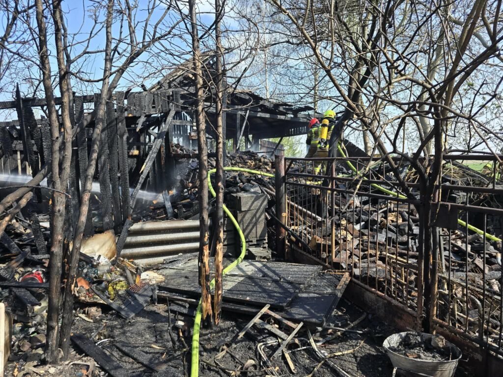 Strażacy gaszą pożar w zniszczonym budynku, otoczonym poparzonymi drzewami i resztkami spalonego materiału.