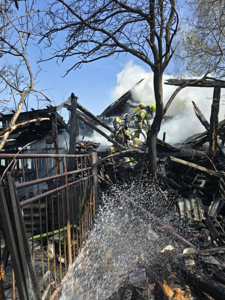 Strażacy gaszą pożar zniszczonego budynku, otoczeni przez dym i popioły, w słoneczny dzień.