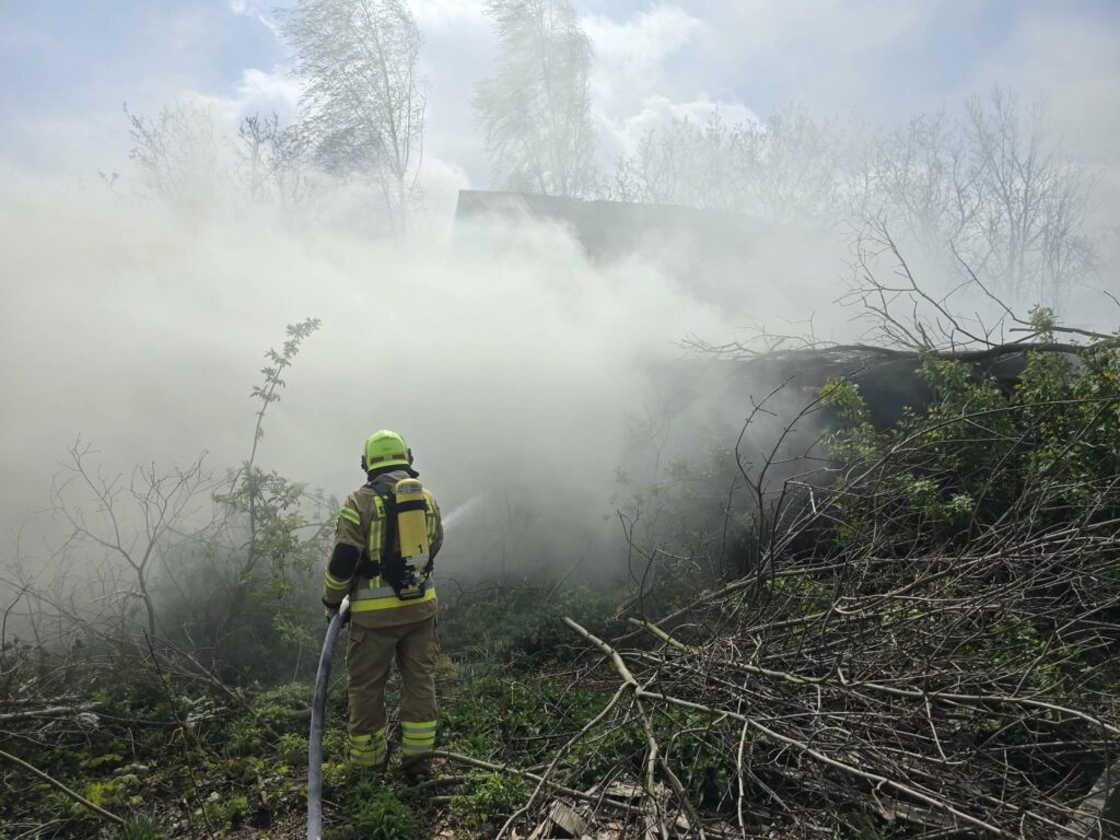 Strażak w hełmie i mundurze, trzymający wąż gaśniczy, stoi w gęstym dymie pożaru w otoczeniu zarośli.