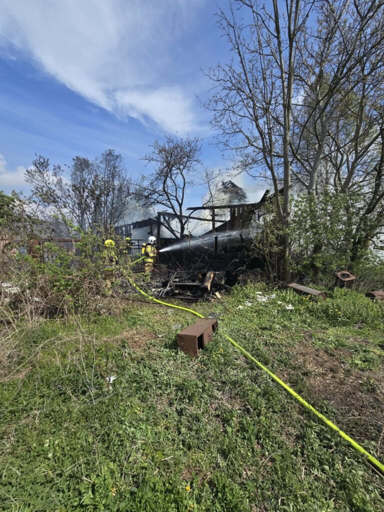 Strażacy gaszą ogień w zniszczonym budynku, otoczonym przez zieloną roślinność i dym unoszący się w powietrzu.