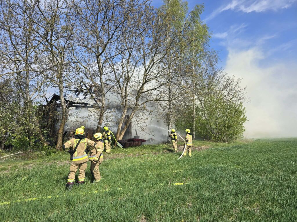 Strażacy w hełmach gaszą pożar budynku w otoczeniu drzew i zielonego pola, w tle widoczny dym.