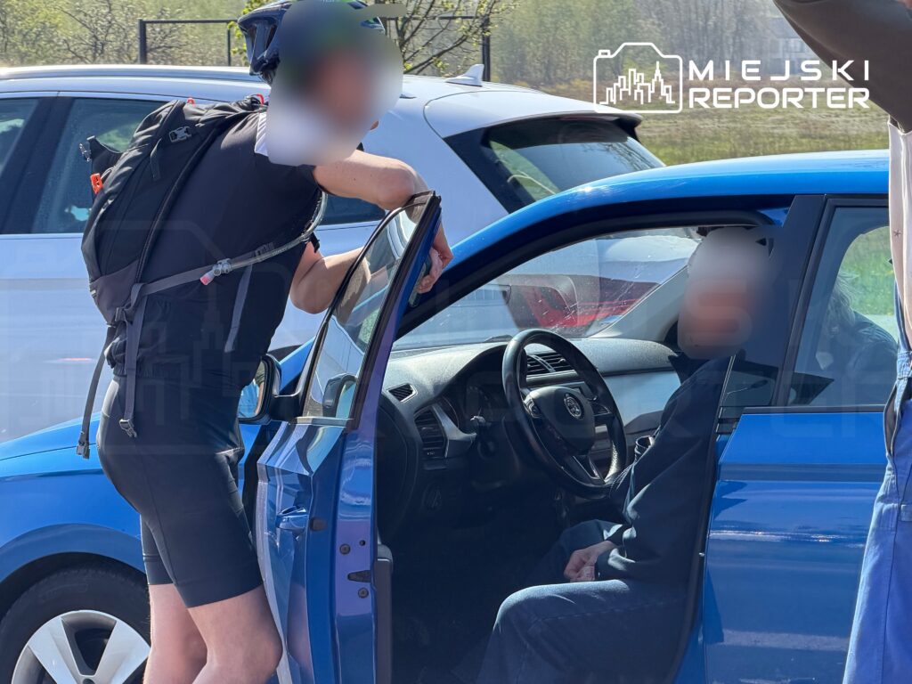 Rowerzysta w sportowym stroju rozmawia z kierowcą siedzącym w niebieskim samochodzie na parkingu.