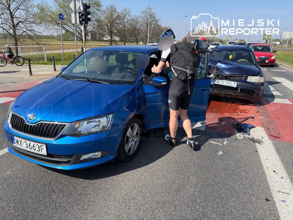 Rowerzysta w kasku rozmawia z kierowcą niebieskiego samochodu osobowego na skrzyżowaniu, obok stoi uszkodzony czarny pojazd.