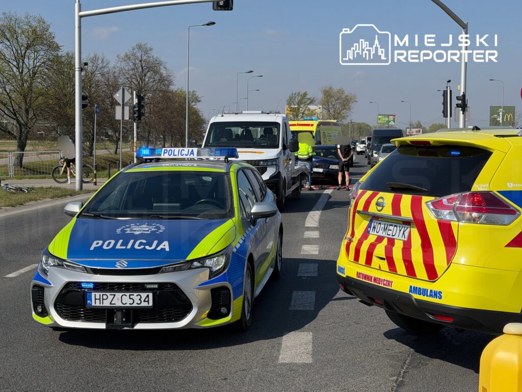 Na skrzyżowaniu stoi radiowóz policji oraz karetka, a obok trwają czynności związane z wypadkiem drogowym.