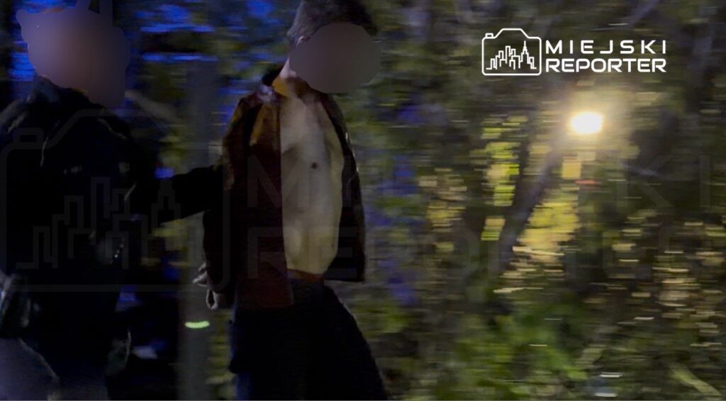 Mężczyzna w brązowej kurtce prowadzony przez funkcjonariusza w oświetlonym parku nocą.