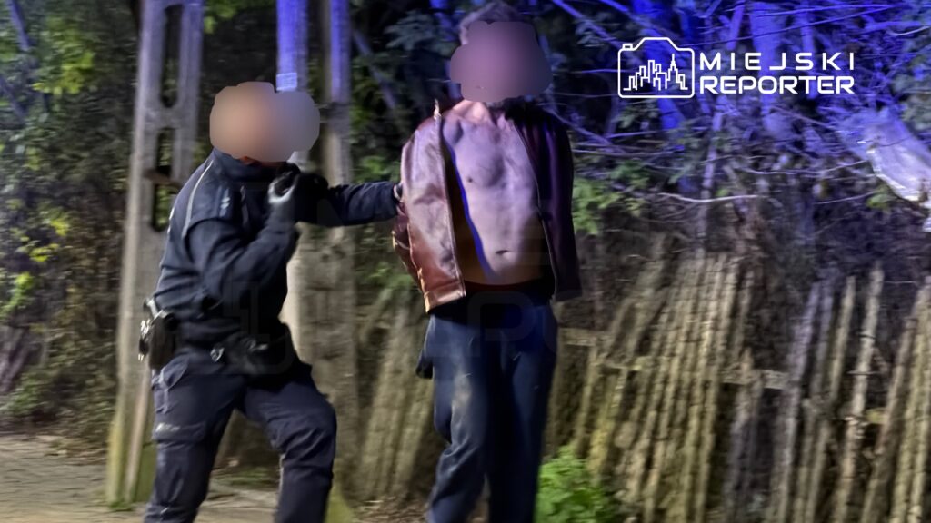 Policjant w mundurze zatrzymuje mężczyznę w skórzanej kurtce w ciemnym otoczeniu, oświetlonym niebieskimi światłami.