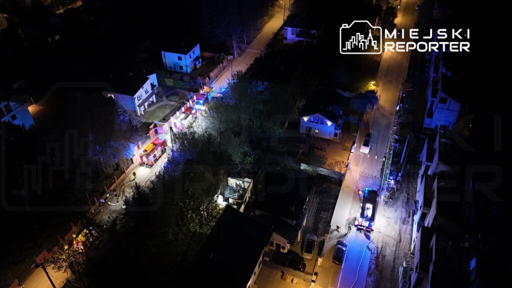 W nocy na ulicy widać pojazdy strażackie z włączonymi sygnałami świetlnymi, a w tle dostrzegalne są domy mieszkalne.