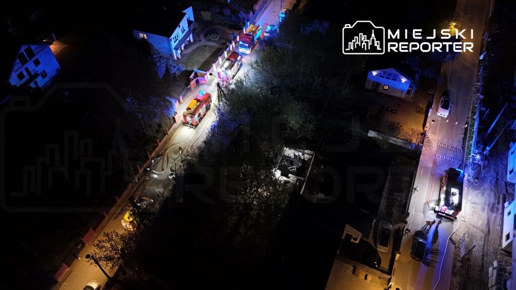 Strażacy z wozami gaśniczymi pracują przy gaszeniu pożaru w zabudowanej okolicy nocą, otoczeni niebieskimi światłami.