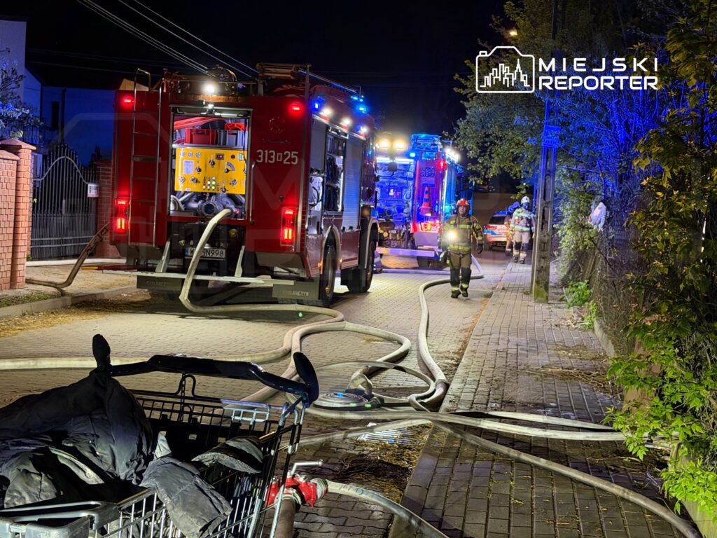 Strażacy w mundurach pracują przy wozie strażackim na oświetlonej ulicy, rozkładając węże gaśnicze.