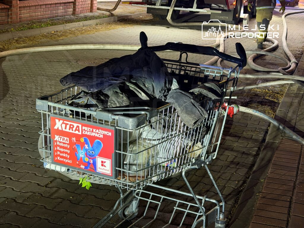 Wózek sklepowy z czarnymi ubraniami leży na chodniku obok węża strażackiego w nocnym otoczeniu.