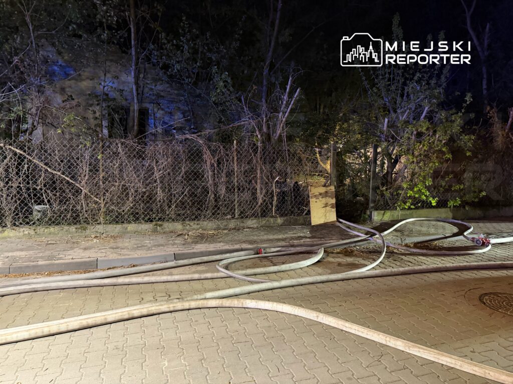 Węże strażackie leżą na chodniku obok ogrodzenia, w otoczeniu zarośli i słabego oświetlenia nocnego.