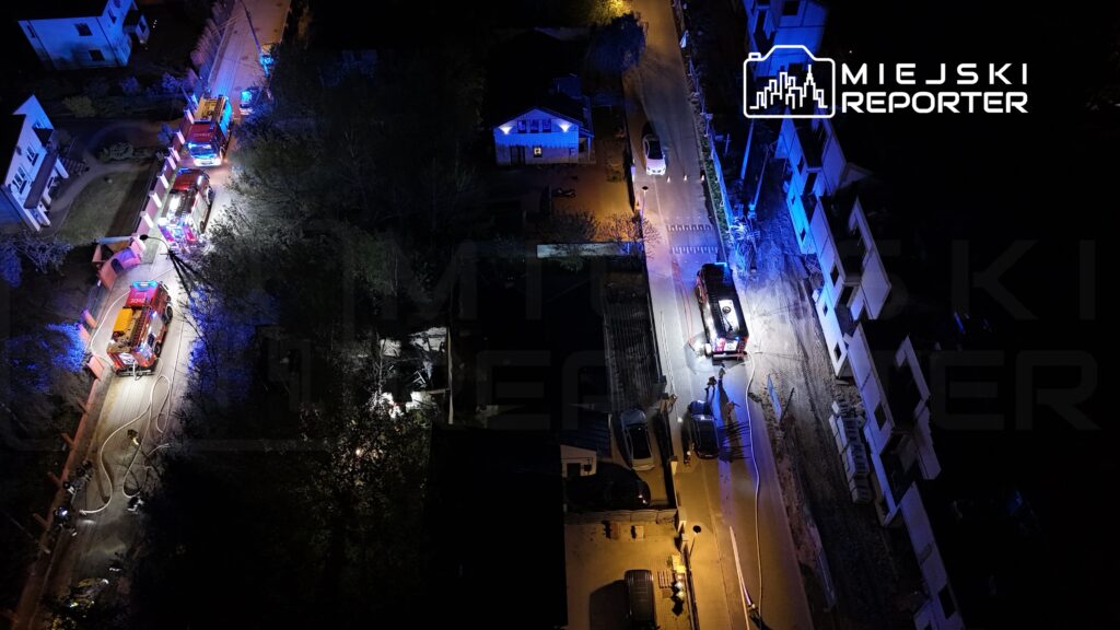 Kilka wozów strażackich z włączonymi syrenami i niebieskimi światłami na oświetlonej ulicy, w nocy.