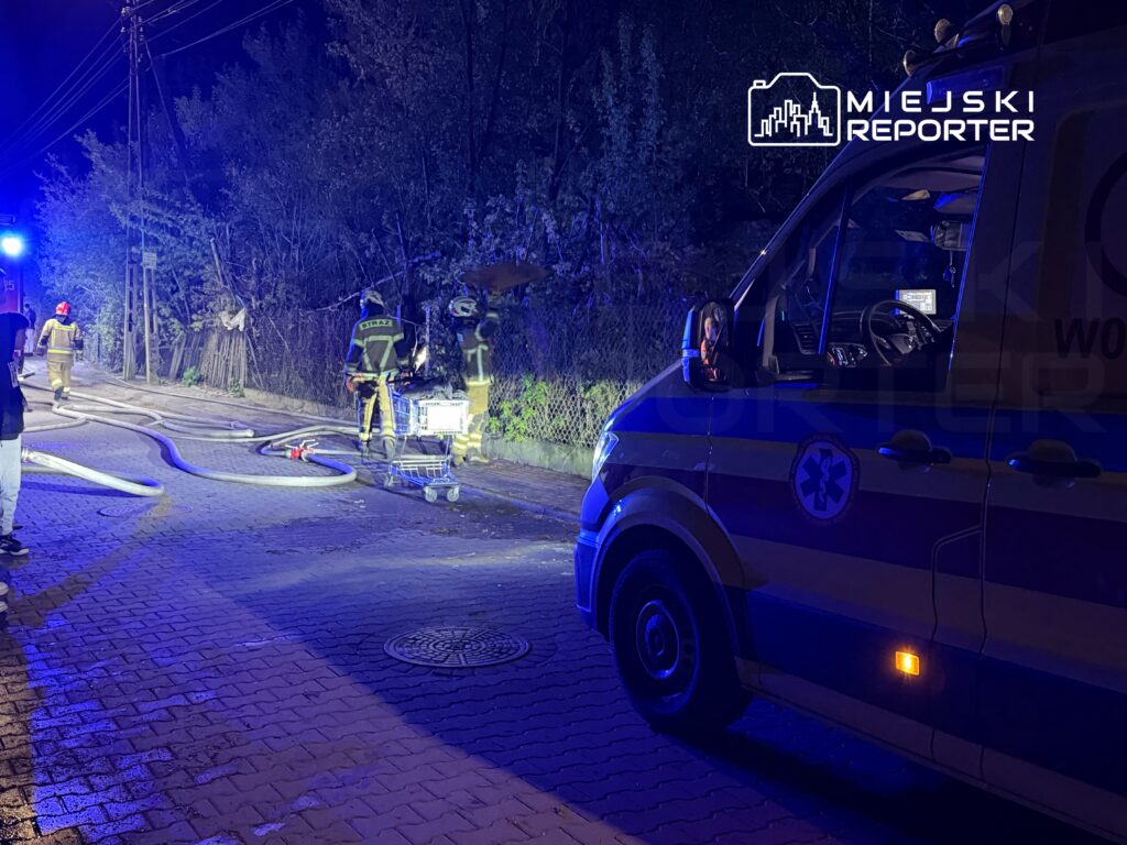 Strażacy w specjalnych strojach pracują przy wężach pożarniczych obok karetki na oświetlonej ulicy.