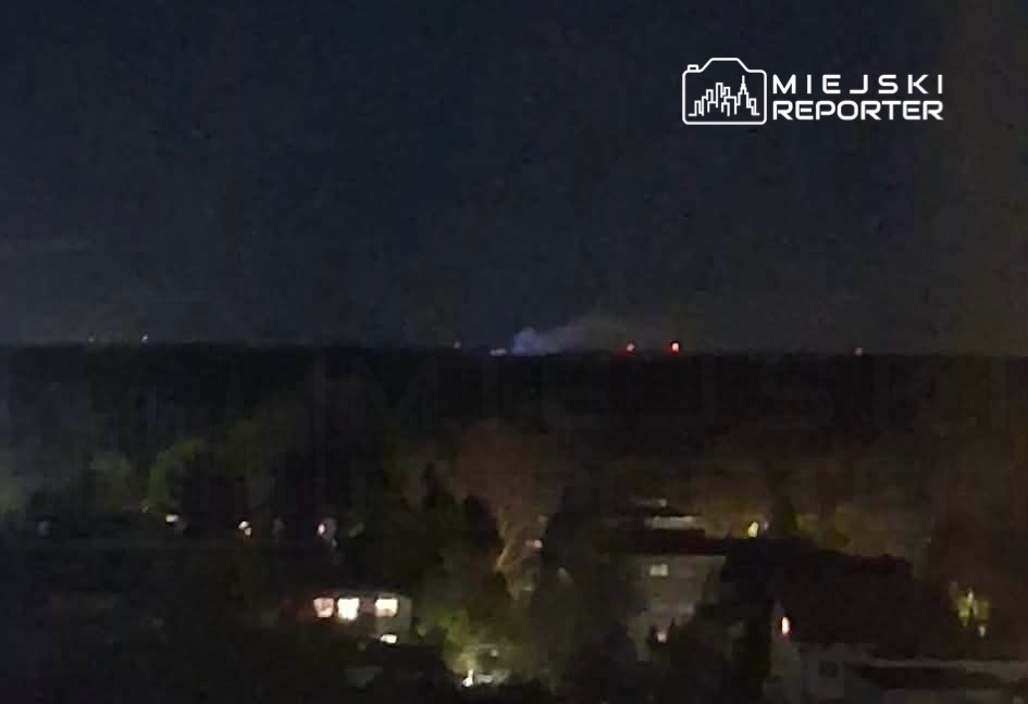 Na horyzoncie widoczne dymiące obiekty w nocy, w tle ciemne niebo i oświetlone domy.