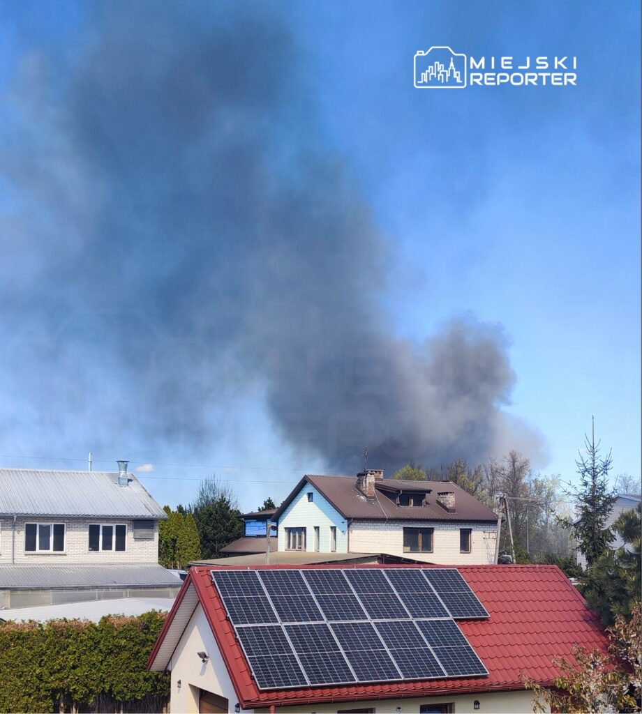 Gęsty, czarny dym unoszący się nad dachami domów w spokojnej okolicy, w tle widoczna instalacja fotowoltaiczna.