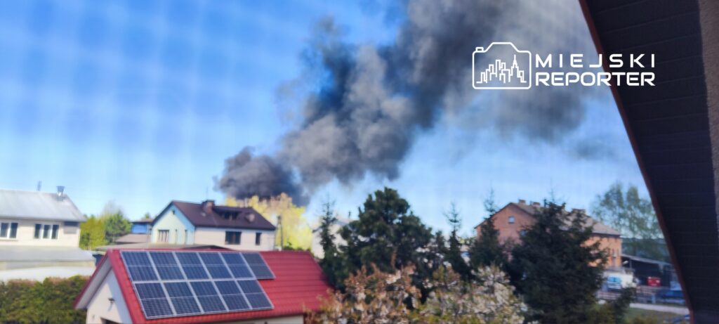 Czarny dym unoszący się z kierunku budynków mieszkalnych w słoneczny dzień. W tle widać dachy domów i drzewa.